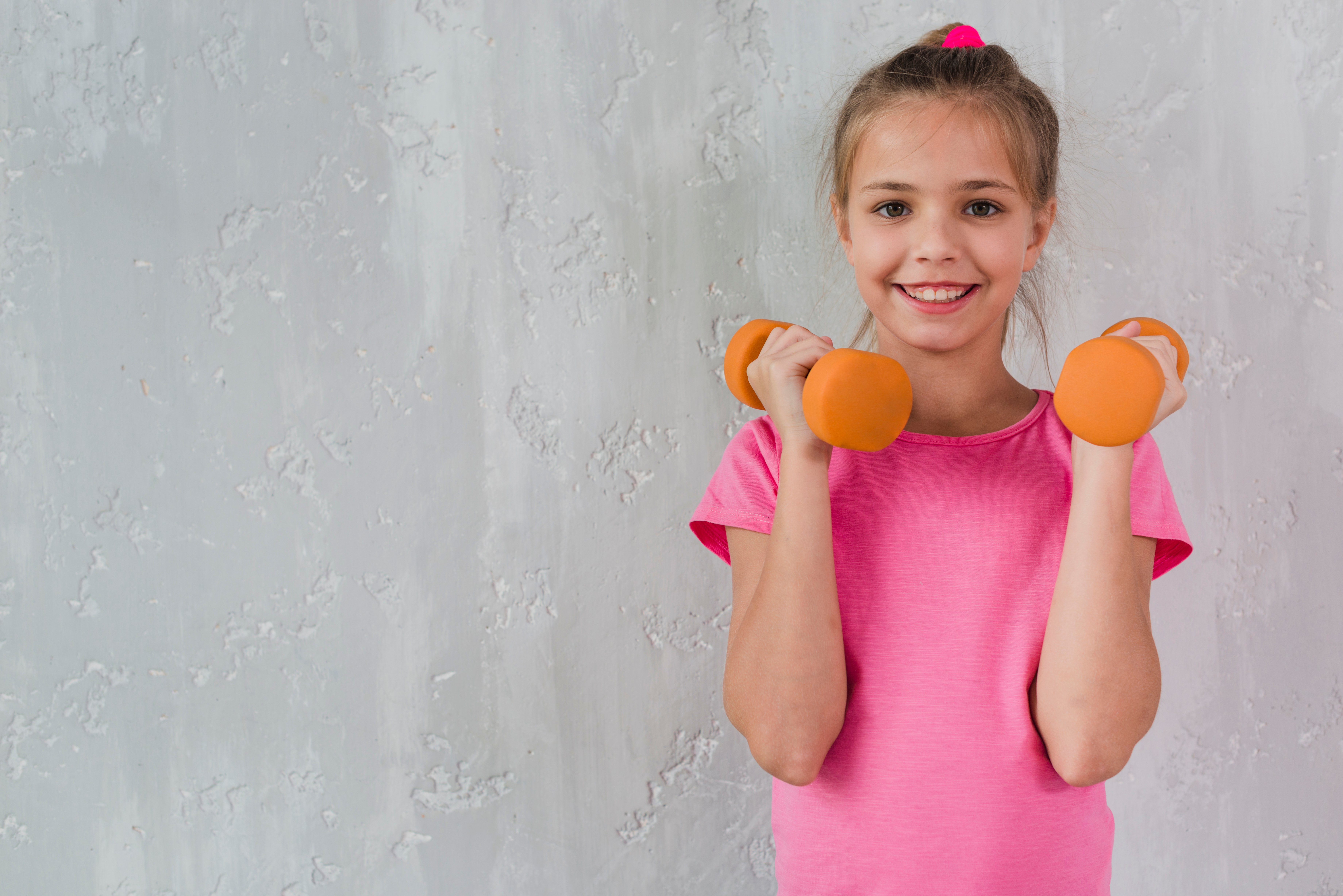 smiling-girl-holding-orange-dumbbell-in-front-of-concrete-wall